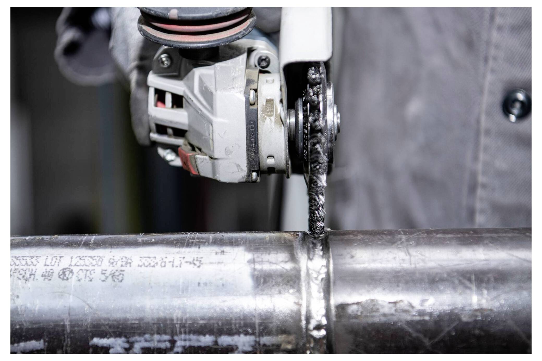 A person uses an angle grinder to cut through a metal pipe, wearing protective gloves. The grinder creates sparks during the cutting process.