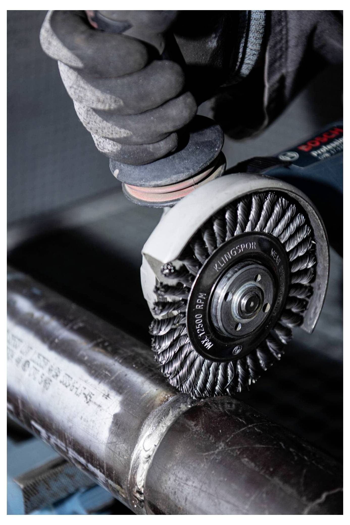 Worker using angle grinder with wire brush attachment to polish metal pipe, wearing protective gloves.