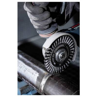 Worker using angle grinder with wire brush attachment to polish metal pipe, wearing protective gloves.