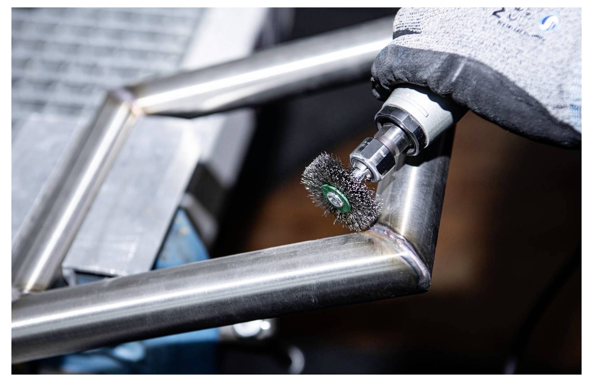 A close-up of a person using a rotary wire brush tool to polish a metal frame corner, enhancing its finish and removing debris.