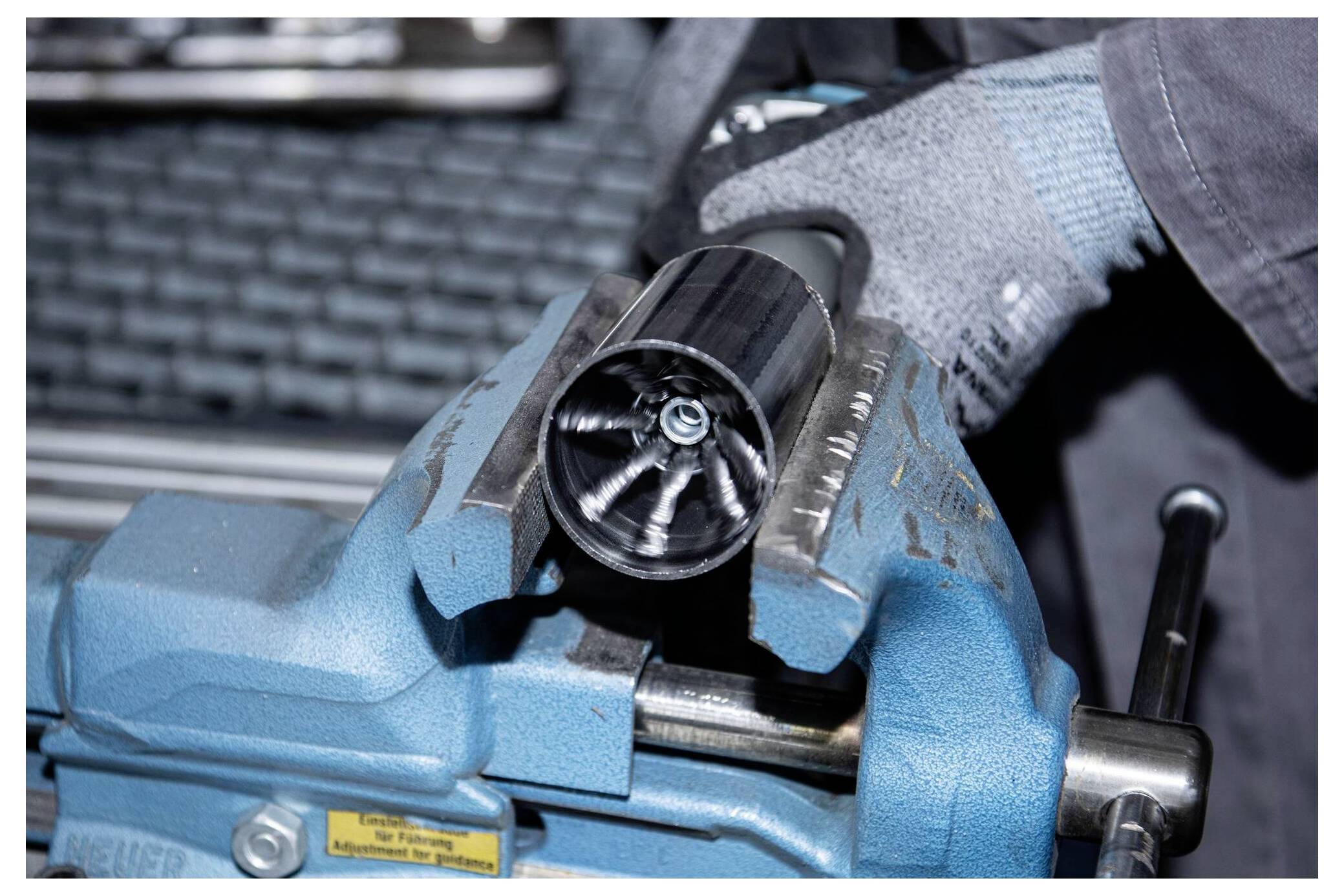 A person wearing gray gloves uses a blue metal vise to secure a cylindrical metal part with a shiny, star-shaped interior.