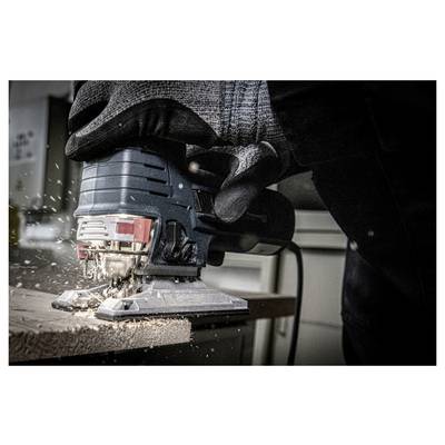A person wearing gloves is using a jigsaw to cut a wooden plank, with wood shavings and sparks flying around.