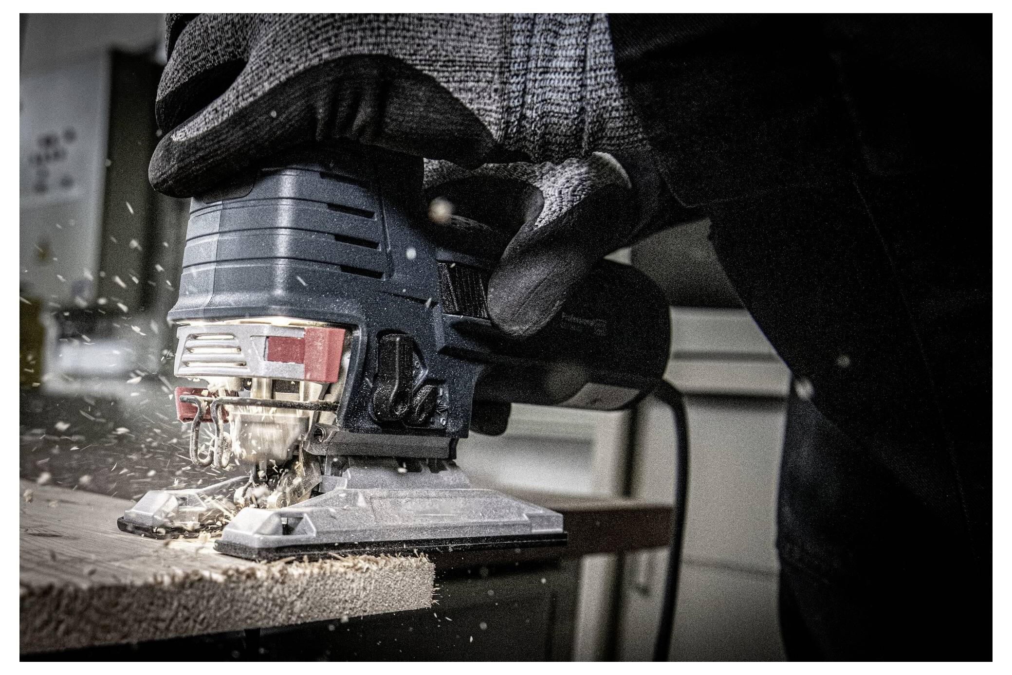A person wearing gloves uses an electric jigsaw to cut through a wooden board, with sawdust scattering in the air.