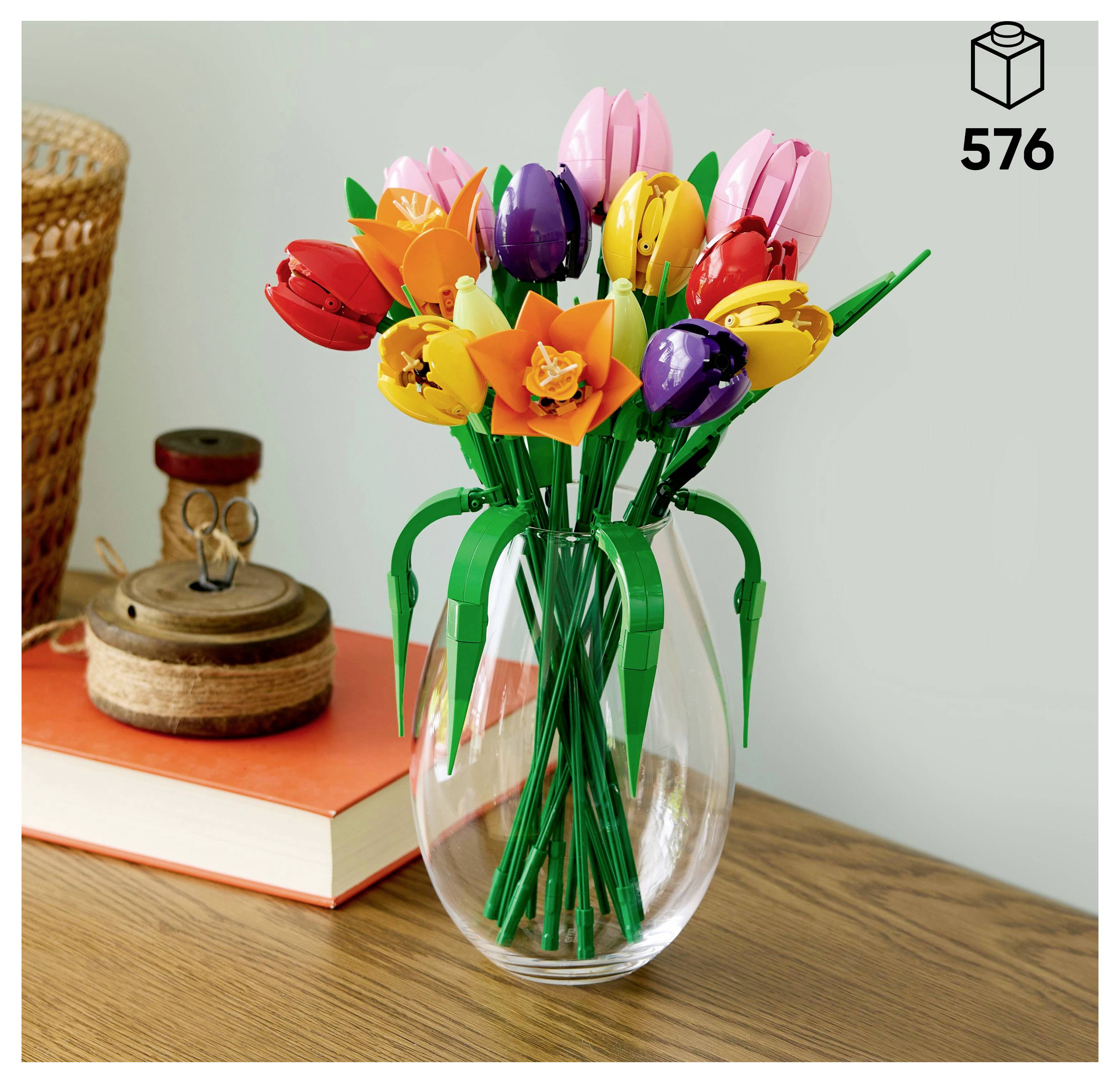 'A bouquet of colorful, LEGO brick flowers in a clear vase, placed on a wooden table beside a book, a wicker basket, and a cork container.'