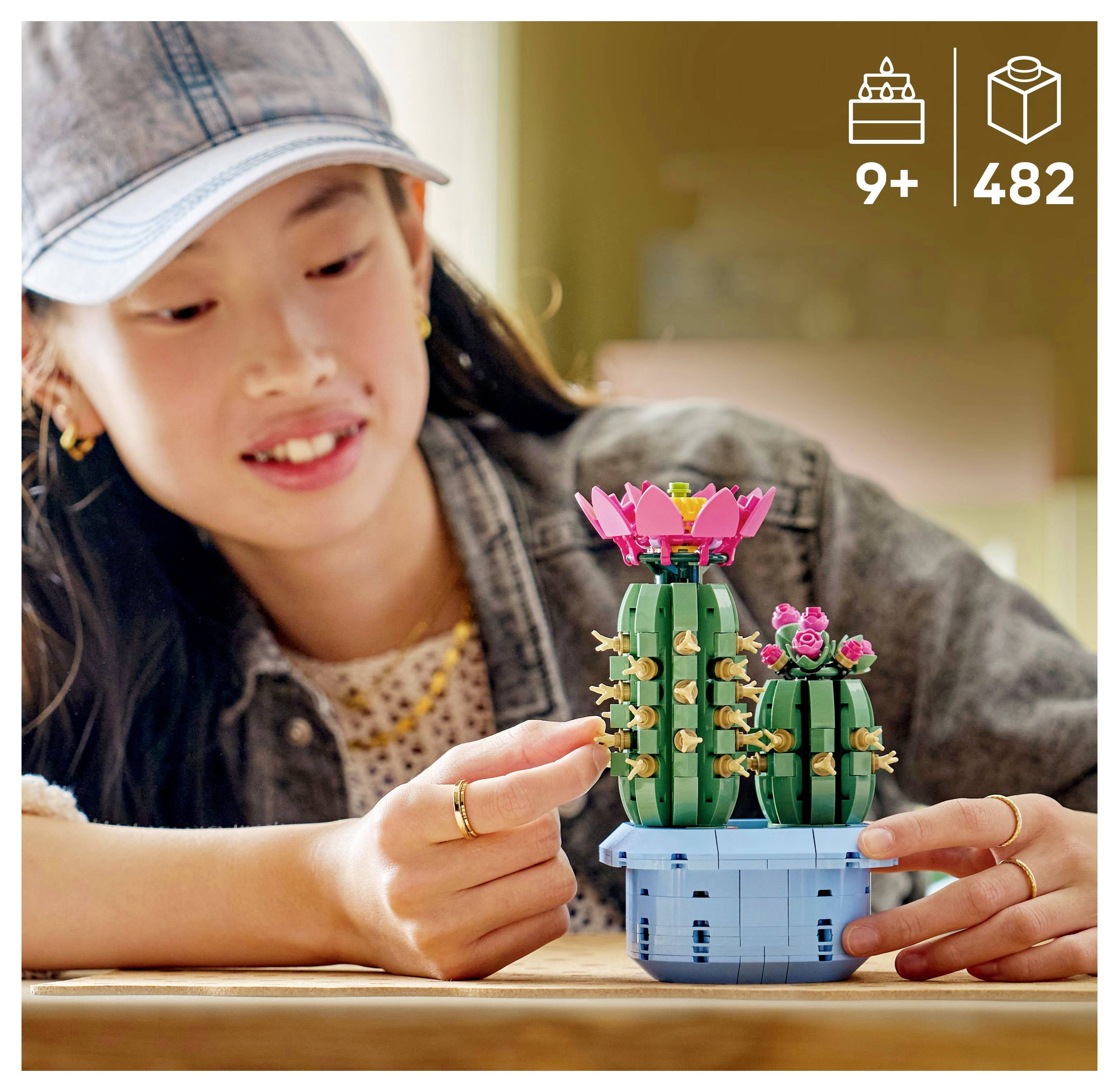A child smiles while assembling a LEGO set of cacti with pink flowers. Packaging shows age recommendation '9+' and '482' pieces.