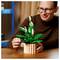 A person assembles a LEGO flower model on a wooden table, surrounded by shelves. The model features green leaves and white blossoms.