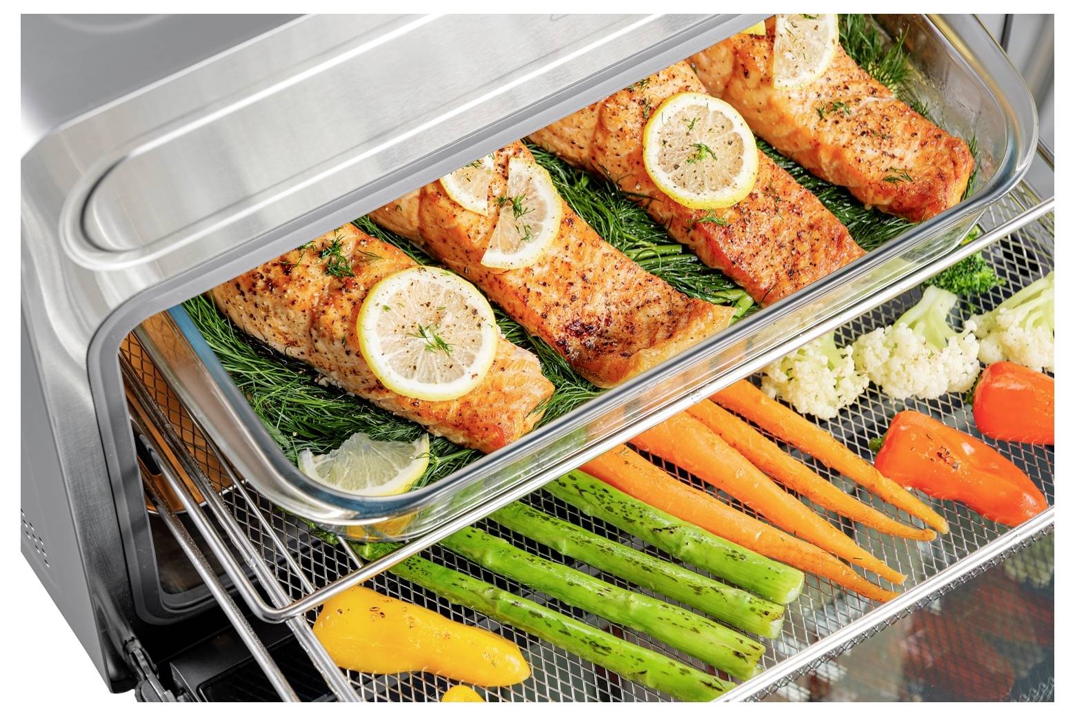 Salmon fillets garnished with lemon and herbs on a tray in an oven, with carrots, asparagus, peppers, and cauliflower on the rack below.
