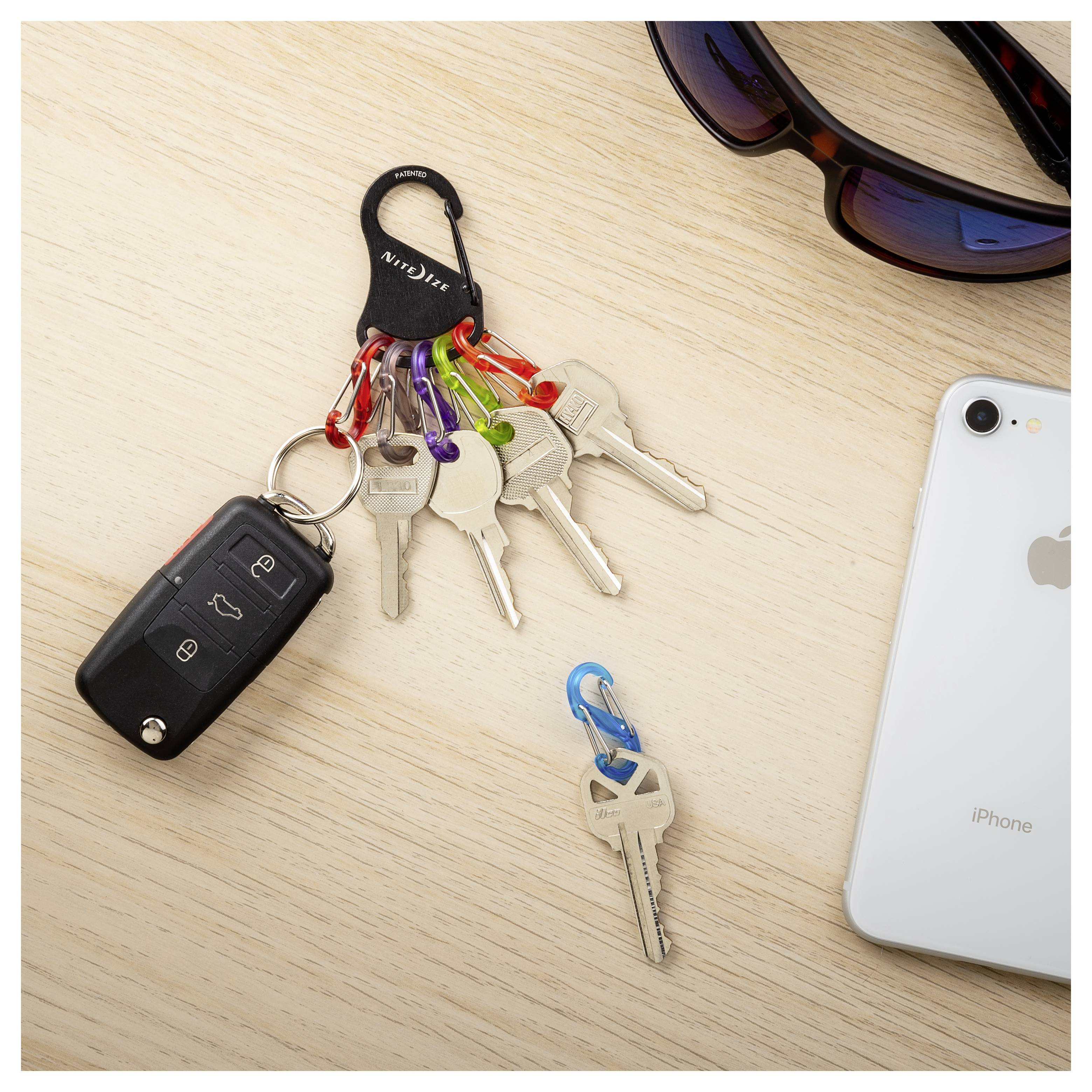 'Set of keys with a black car key remote, colorful keychains on a light wood surface. Sunglasses and a smartphone nearby.'
