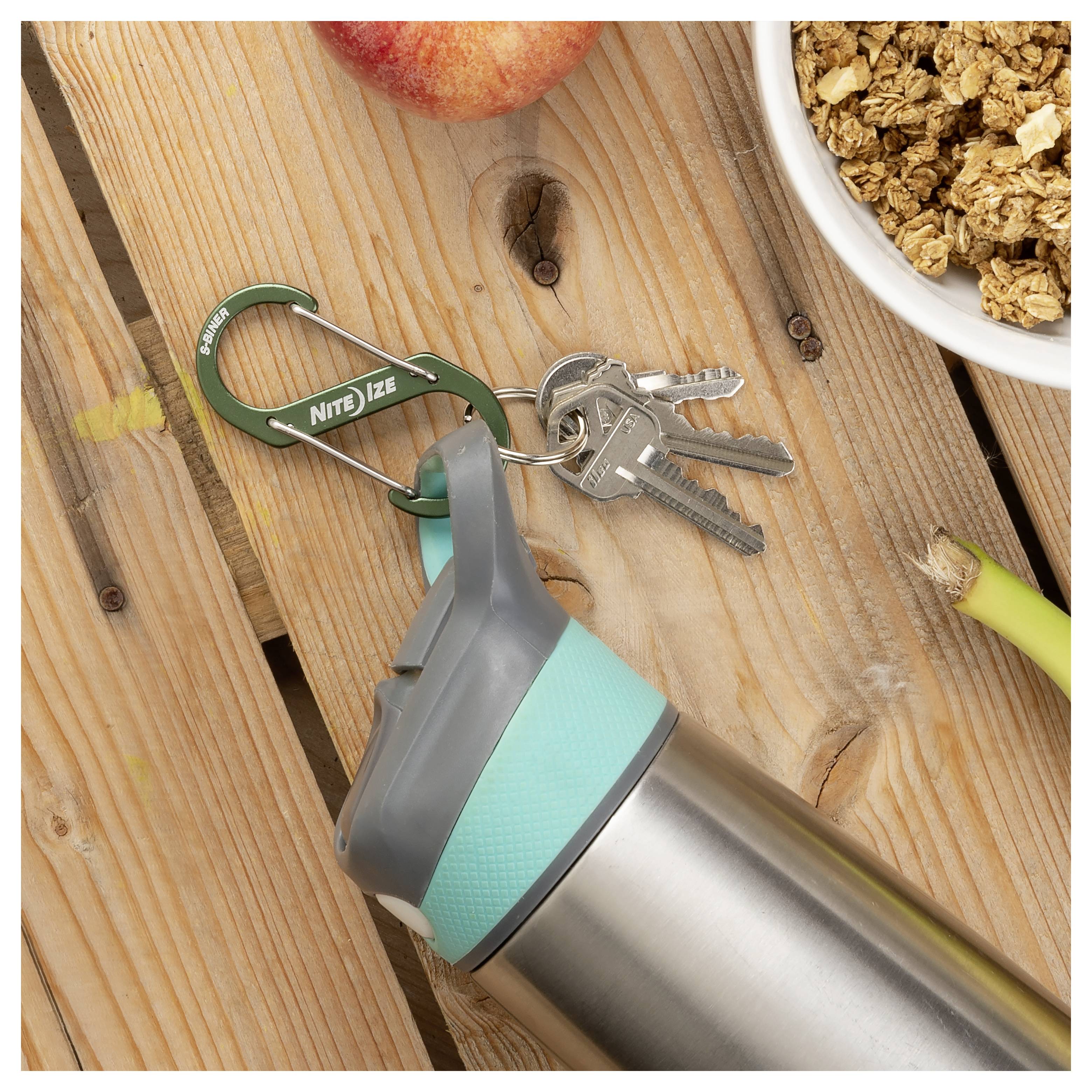 A reusable water bottle with a teal lid is placed on a wooden table next to a set of keys attached to a green carabiner, an apple, and a bowl of granola.