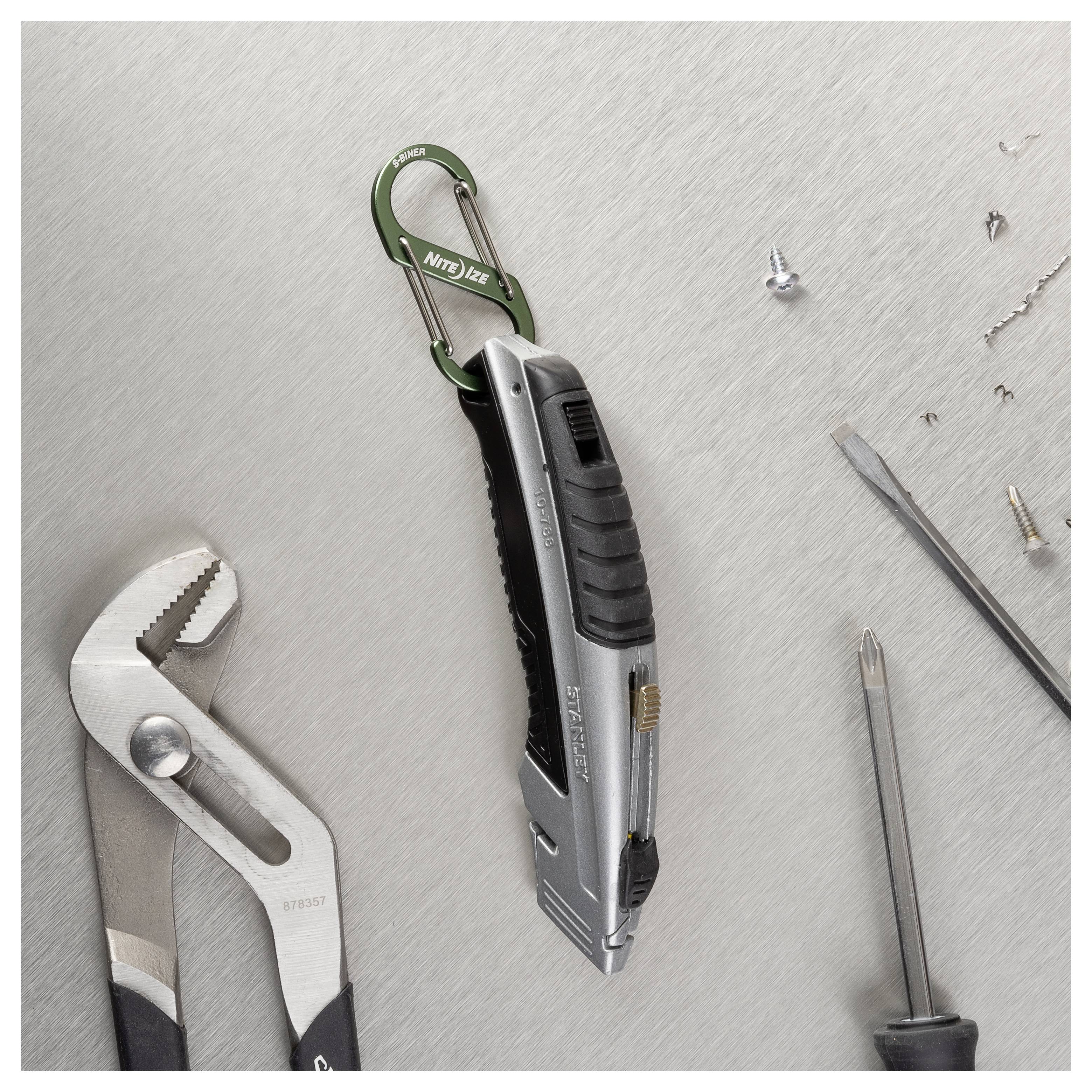 Utility knife on a metal surface, attached to a green carabiner. Surrounding are pliers, a screwdriver, and loose screws.