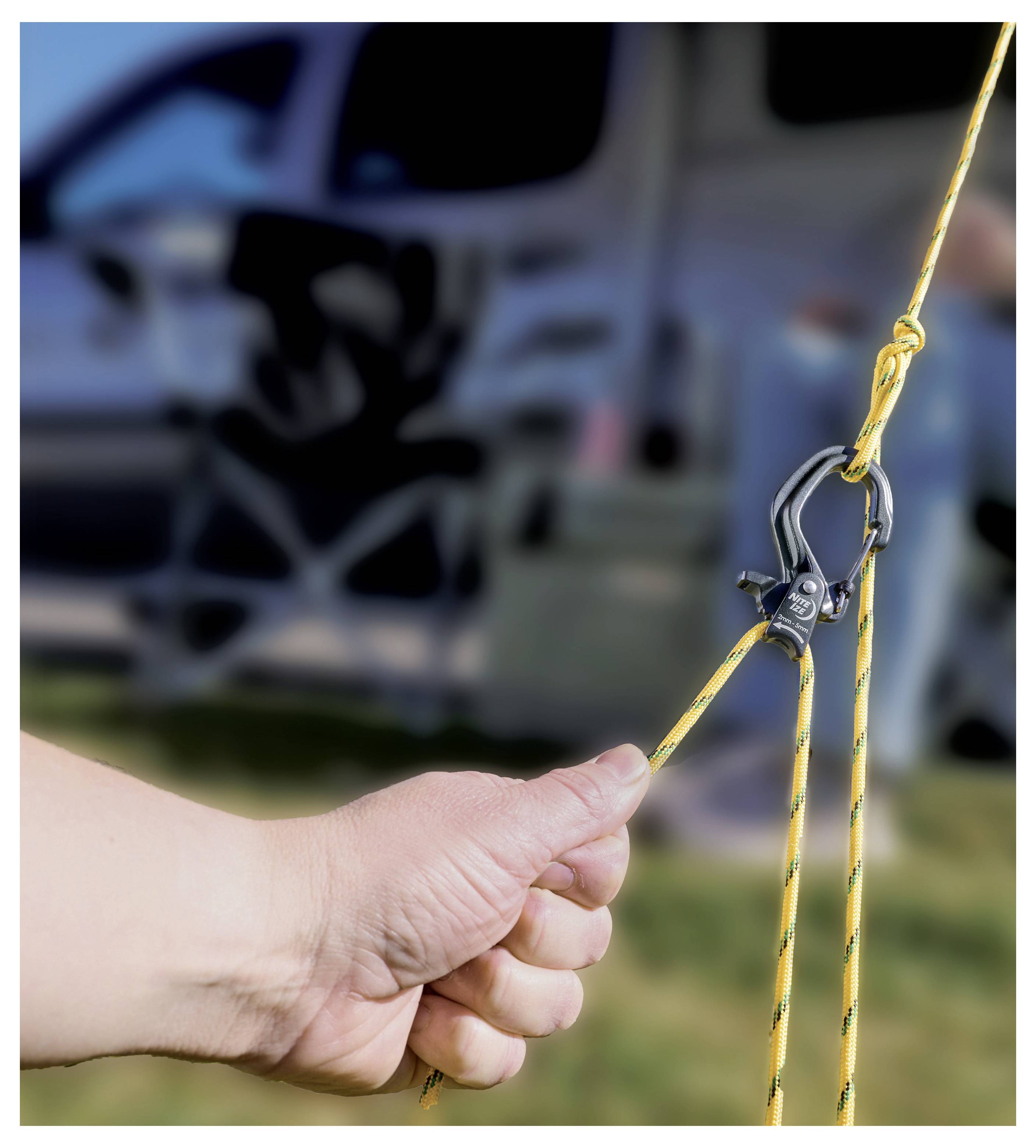 A hand adjusts a yellow rope with a metal clip, likely setting up or adjusting tension. A blurred camping scene is in the background.