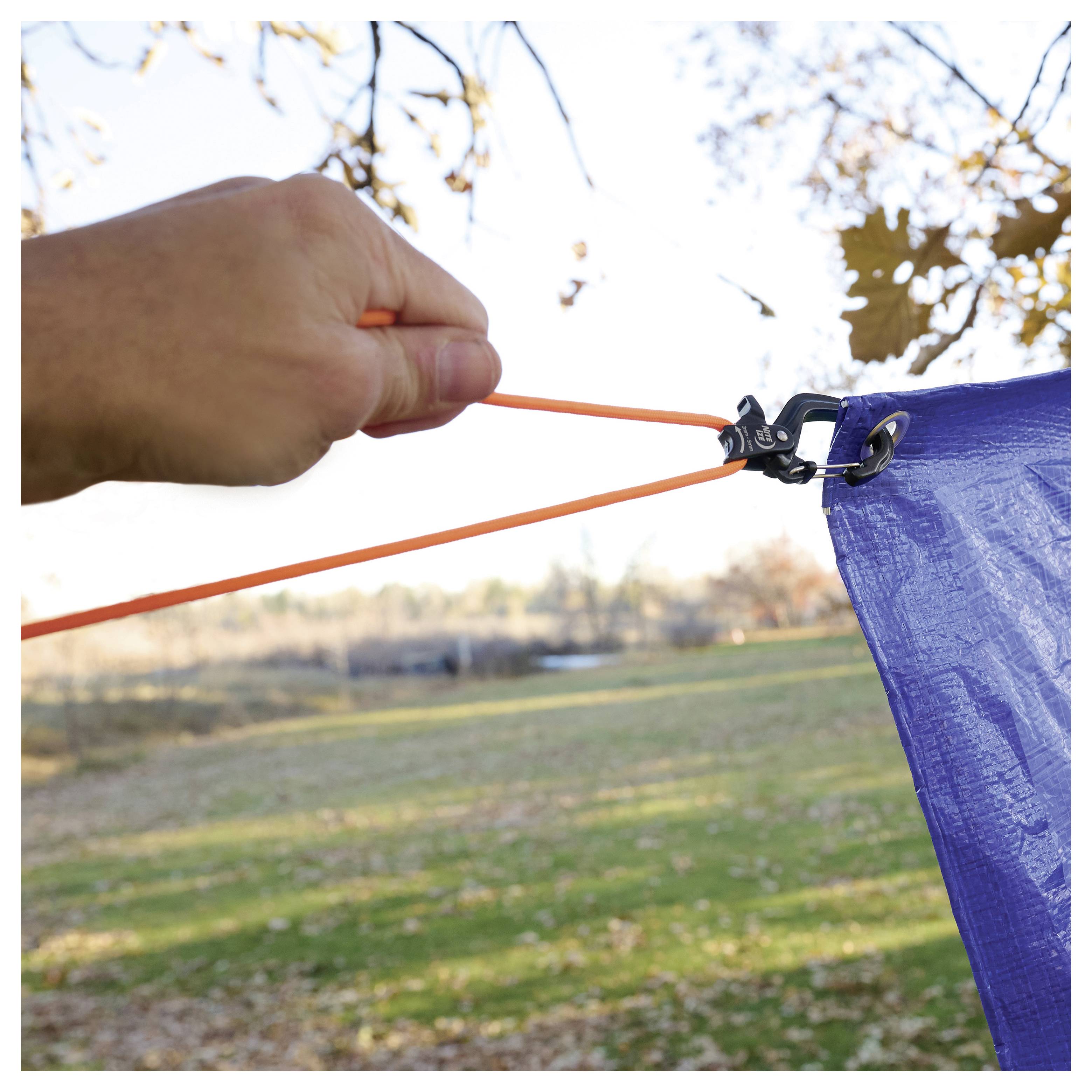 A hand is shown pulling an orange cord to tighten a blue tarp, securing it over a hitch, with an autumn landscape in the background.