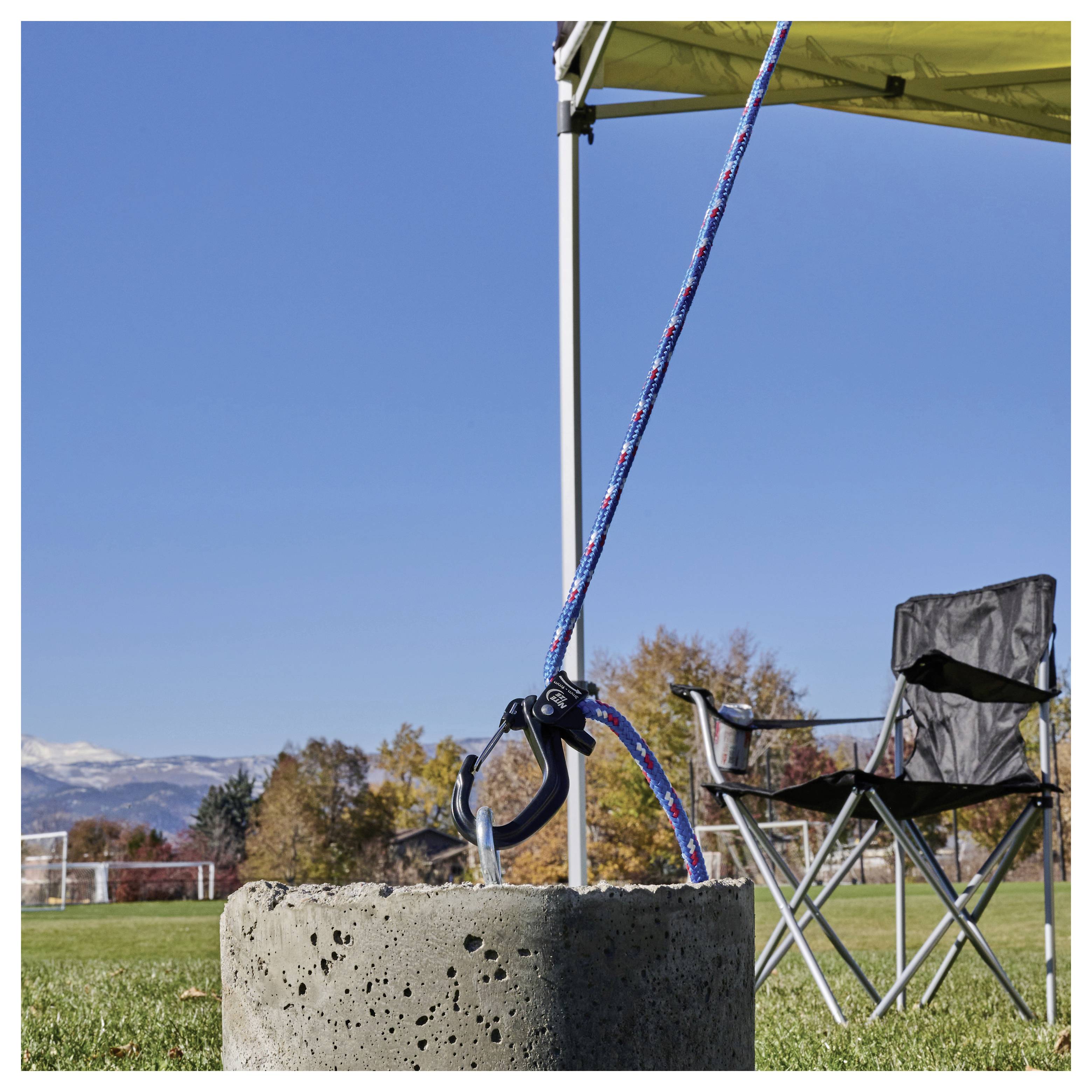 A sturdy concrete anchor holding a tent in place with a blue and red rope, set in a grassy field with autumn trees and mountains in the background.