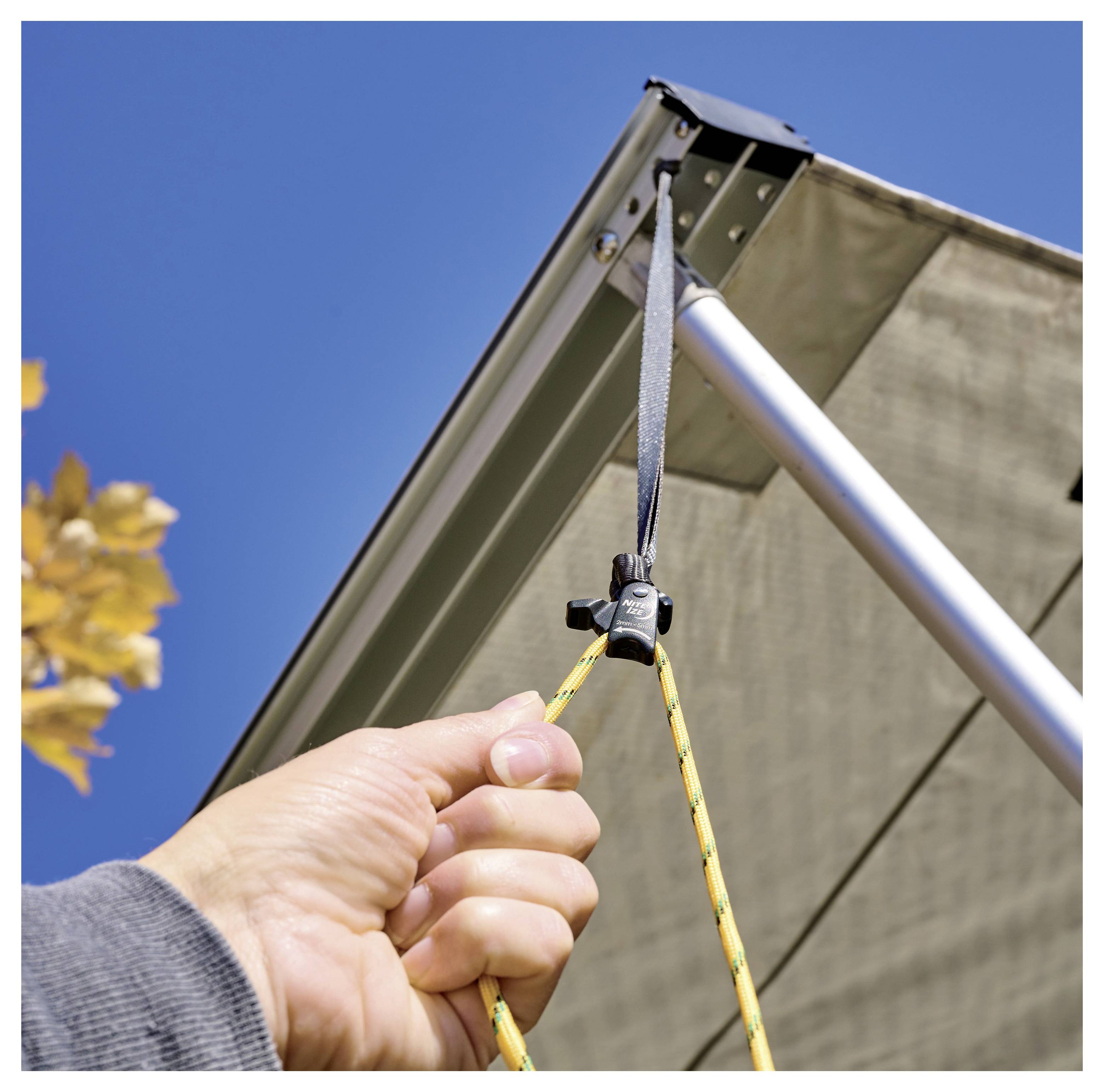 A hand adjusts the tension on a rope attached to a shade awning, with a clear blue sky and a hint of foliage in the background.