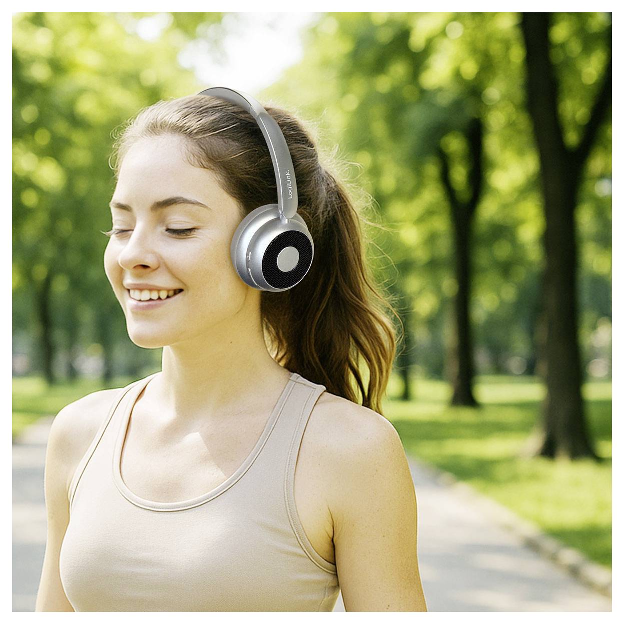 A woman with headphones smiles while walking in a sunny park lined with trees, wearing a beige tank top, suggesting a relaxed mood.