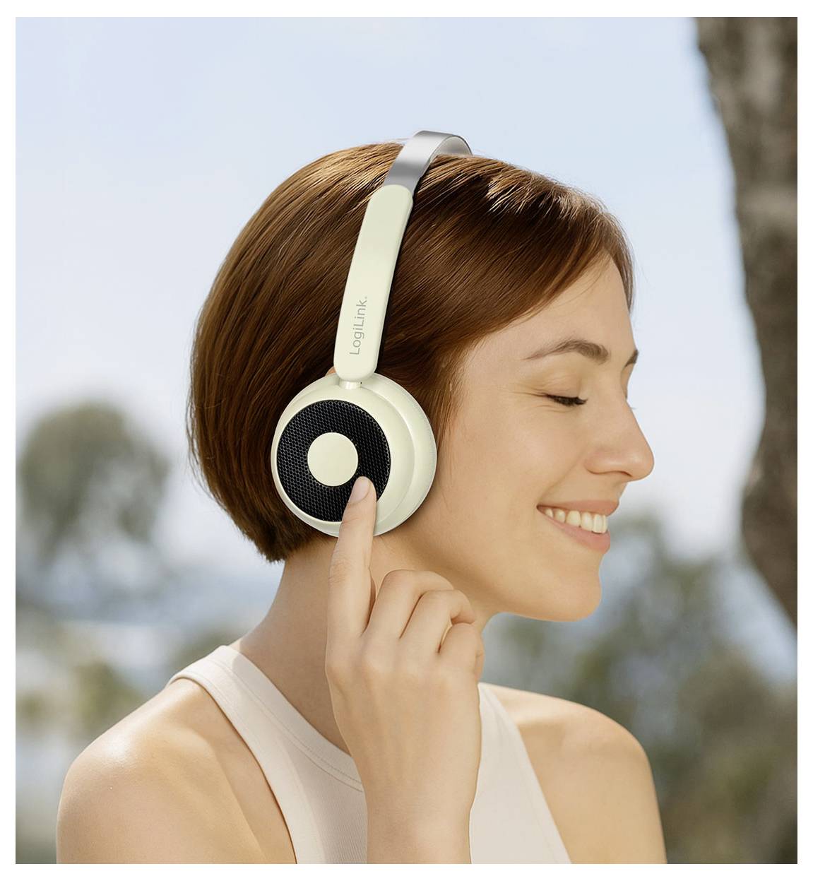 A smiling person with short brown hair wears over-ear headphones outdoors, enjoying music or sound, with trees and blue sky in the background.