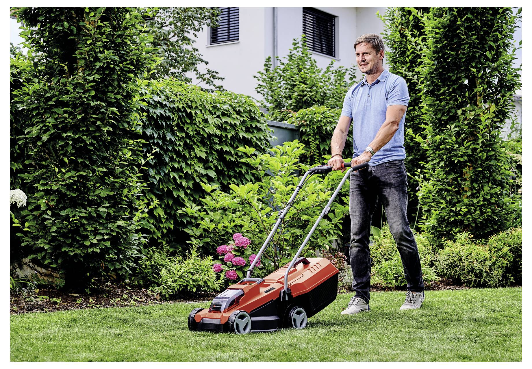 A man is pushing a lawnmower on a grassy lawn. He is outdoors, surrounded by greenery and a flowering bush, with a house in the background.