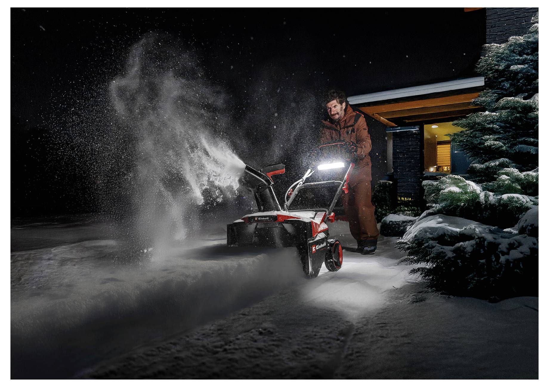 A person wearing warm clothing clears snow from a driveway at night using a snow blower, with a lit house in the background.