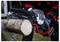 A person wearing protective gloves cuts a log with a small chainsaw outdoors. Sawdust flies as the chainsaw slices through the wood.