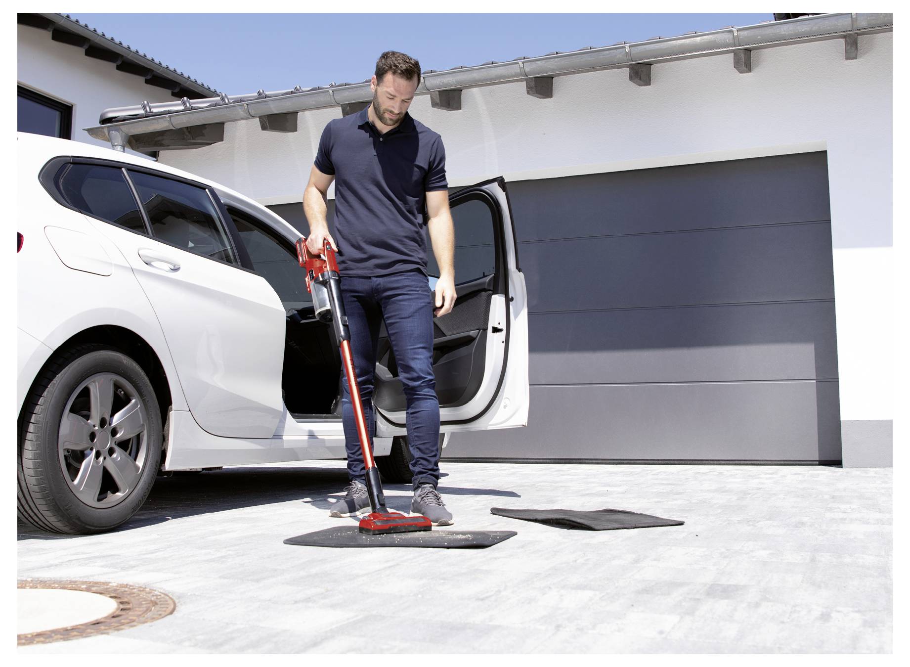 A man vacuums car mats outside next to a white car parked in front of a garage.