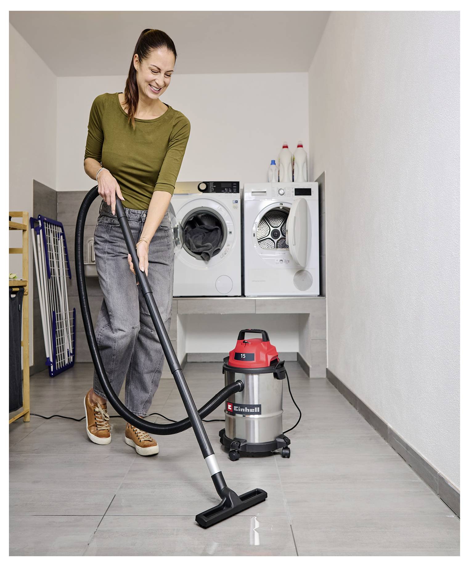A woman uses a vacuum cleaner on a tiled floor in a laundry room. Behind her are washers, dryers, and a drying rack.
