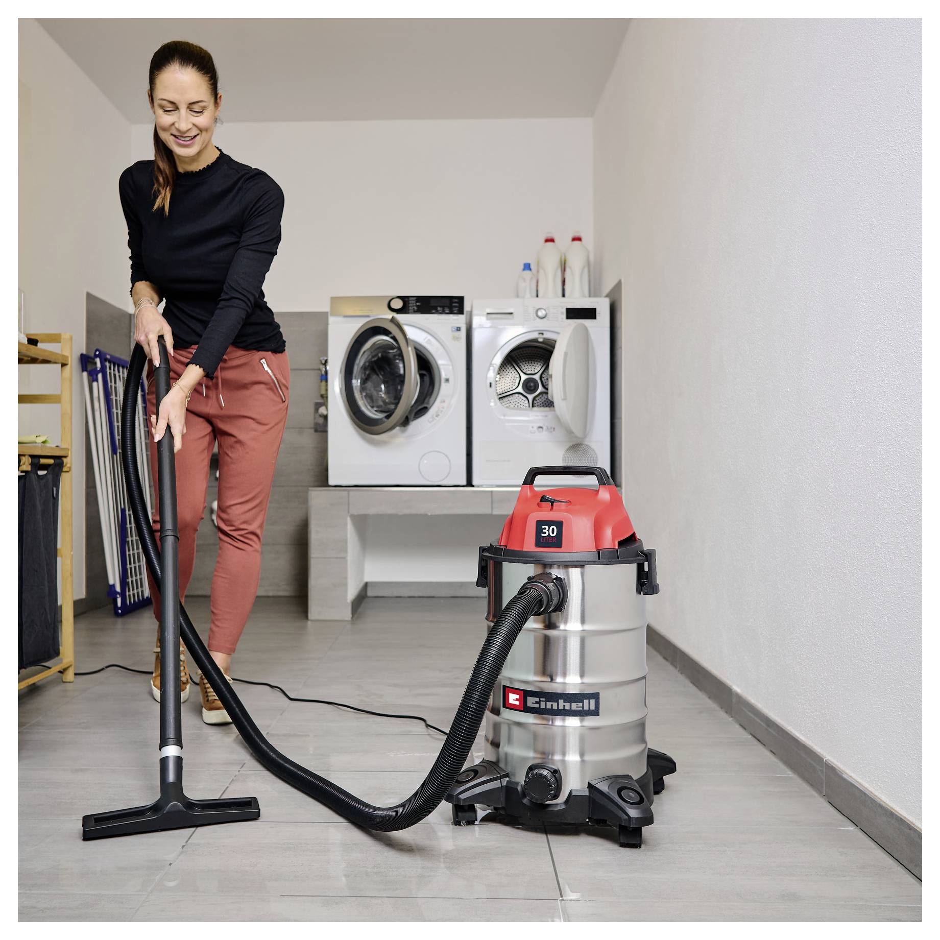A woman vacuuming a room with a wet/dry vacuum cleaner; a washer and dryer are in the background.