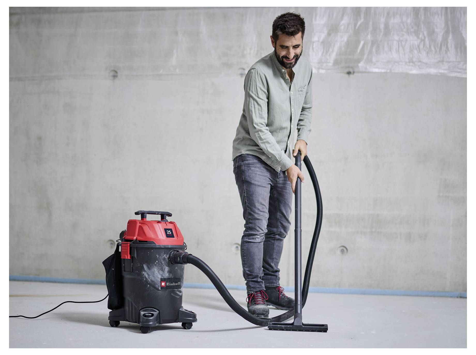 A person uses a red and black wet-dry vacuum cleaner on a bare floor in a concrete-walled room.