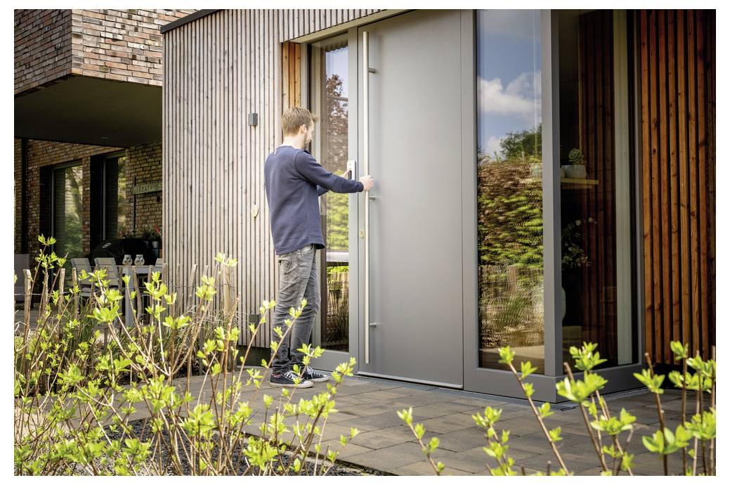 A person opens a modern, sleek, gray front door of a contemporary house with wood and brick exterior, plants visible in the foreground.