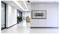 A modern, bright office hallway with large windows, a reception desk in the background, and a wall-mounted TV bracket without a screen.