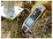 A person uses a handheld electromagnetic field strength meter near a metal post base outdoors, checking signal readings on the display.