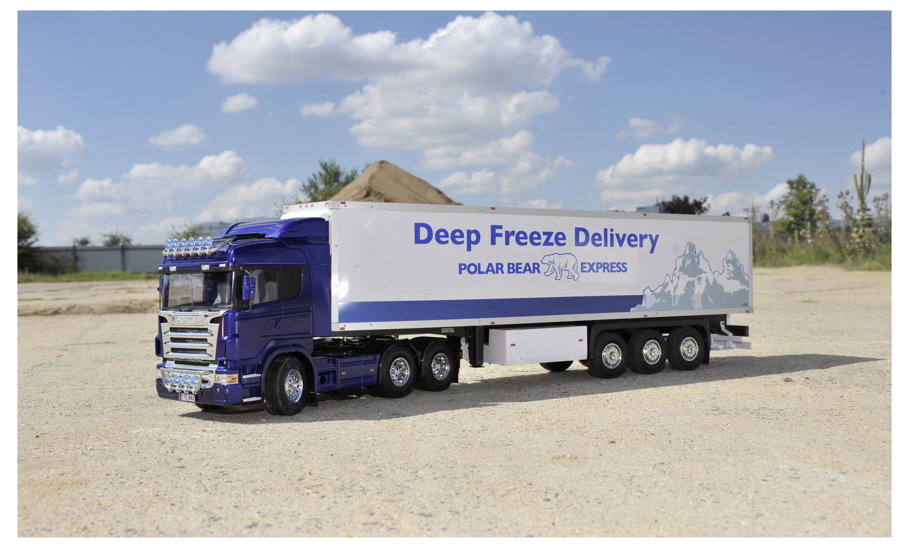 A blue and white truck with 'Deep Freeze Delivery: Polar Bear Express' on the side parked on a gravel lot under a partly cloudy sky.