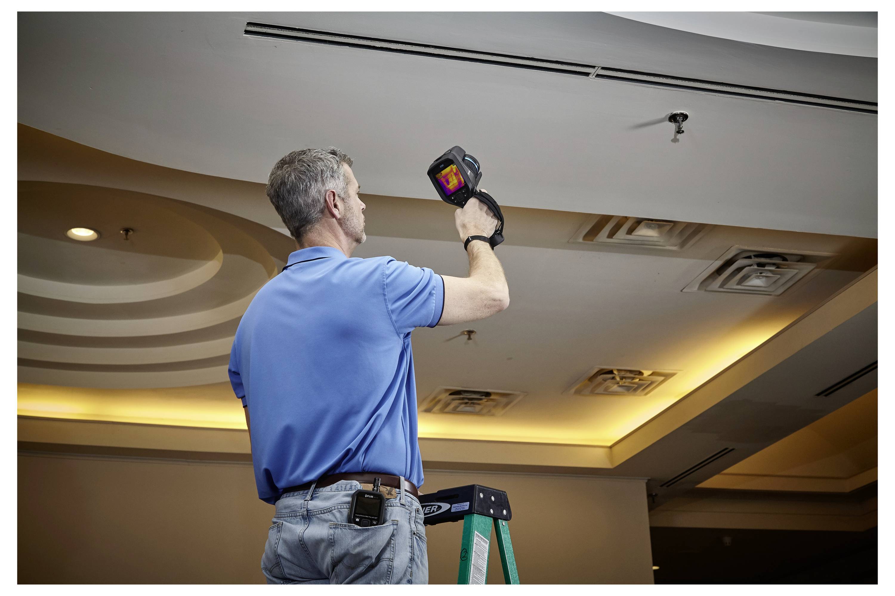 A person in a blue shirt stands on a ladder using a thermal imaging camera to inspect a ceiling, focusing on HVAC vents.