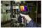 A person holds a thermal imaging camera displaying a color-coded heat map, indicating temperature variations on machinery in a workshop.