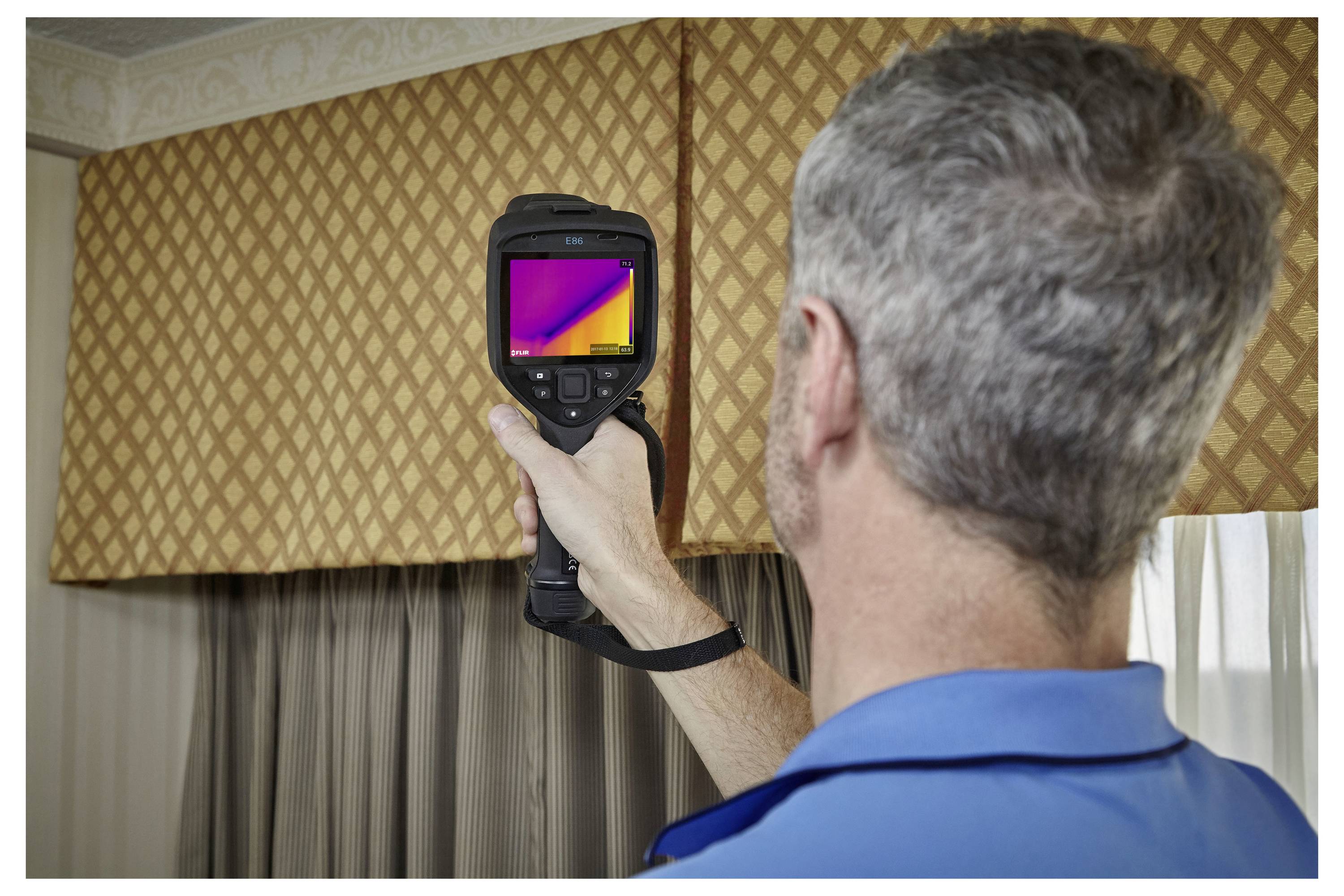 A person using a thermal camera to inspect a room with patterned curtains, likely checking for heat loss or insulation issues.