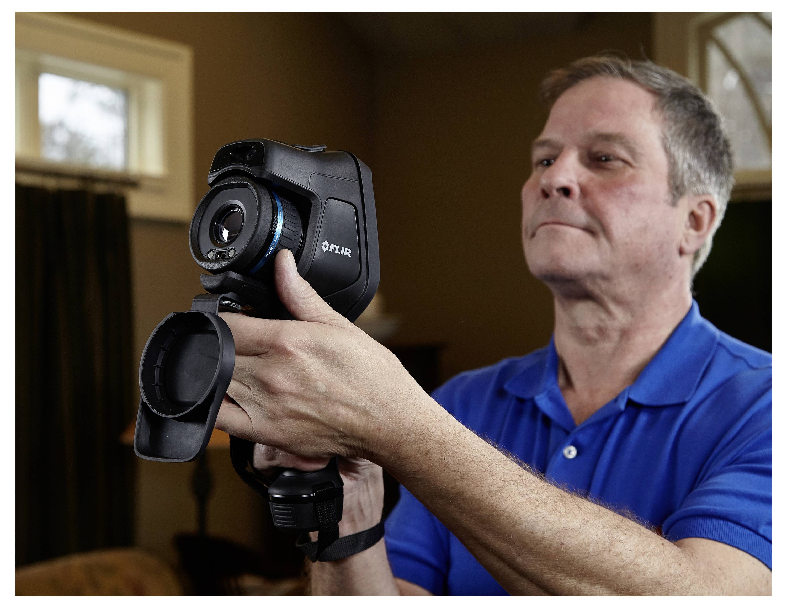 A person in a blue shirt holds a thermal imaging camera indoors, focusing carefully on the screen.
