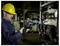 A technician wearing a yellow hard hat uses a thermal imaging camera to inspect electrical panels in an industrial setting.