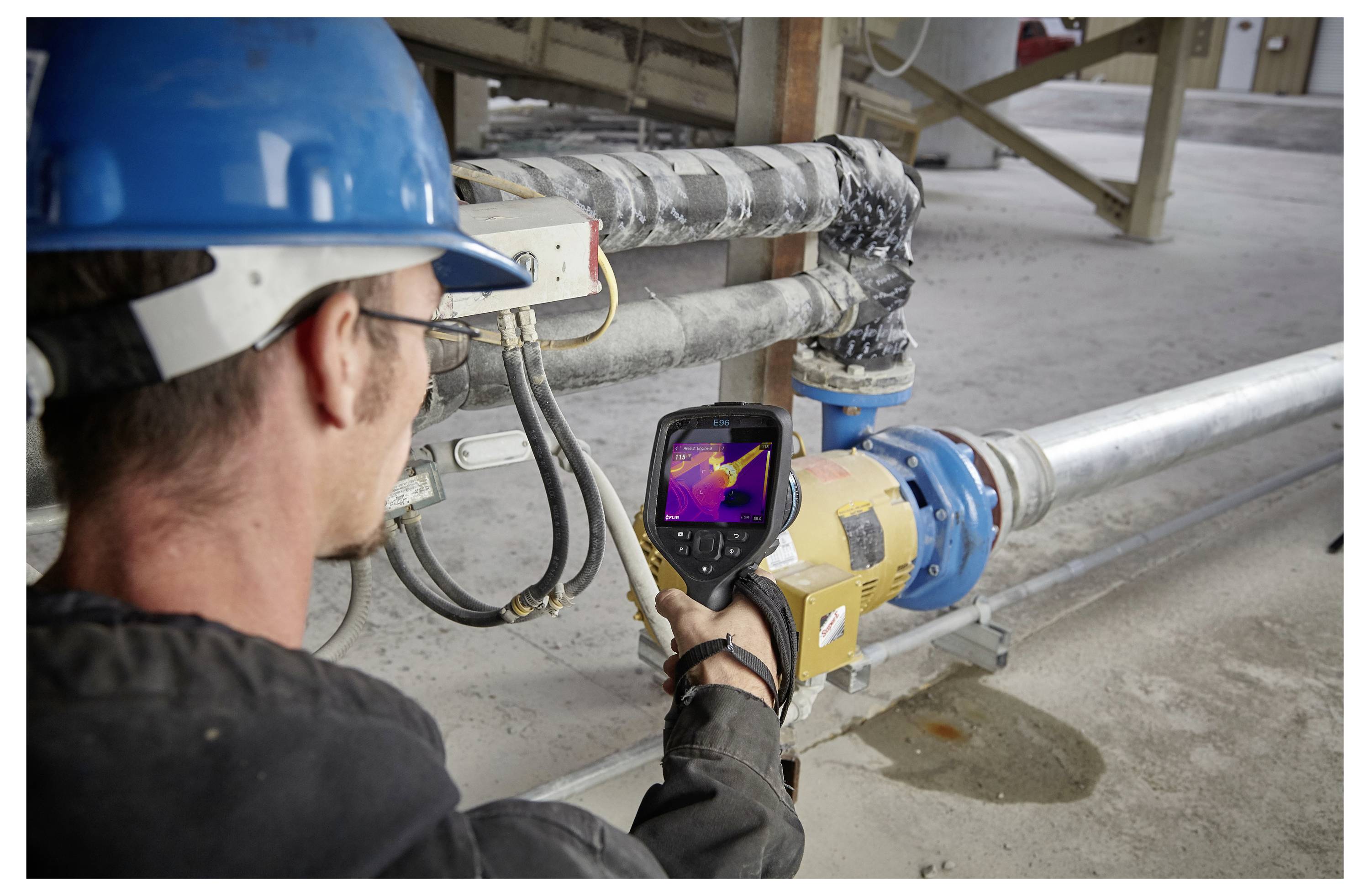 A person wearing a blue hard hat uses a handheld thermal imaging device to inspect pipes in an industrial setting.