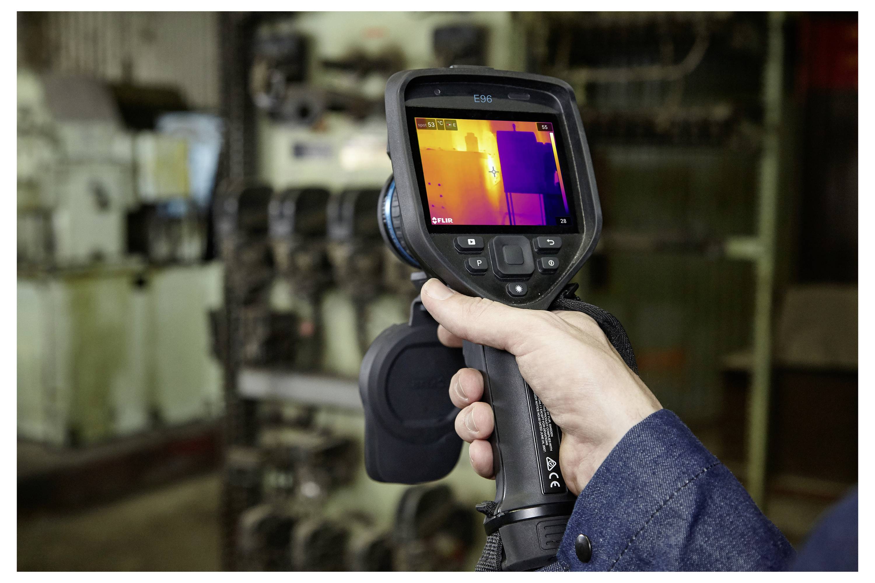 A person holds a thermal imaging camera, displaying a temperature gradient on its screen, likely used for detecting heat variations in machinery.