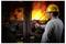 A worker in a hard hat uses a thermal imaging camera to monitor equipment near intense flames in an industrial setting.