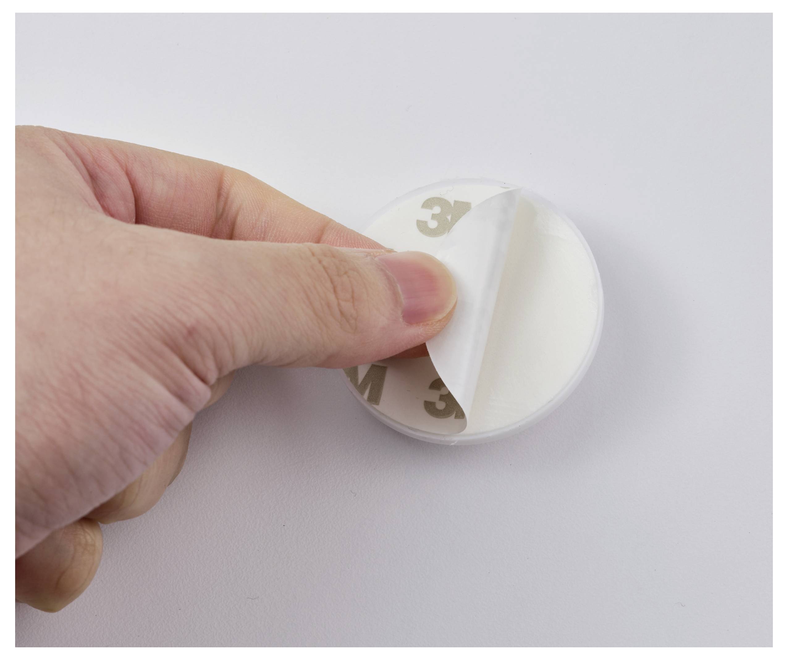 A hand peels the backing off a round adhesive pad, revealing its sticky side for mounting or fixing objects.