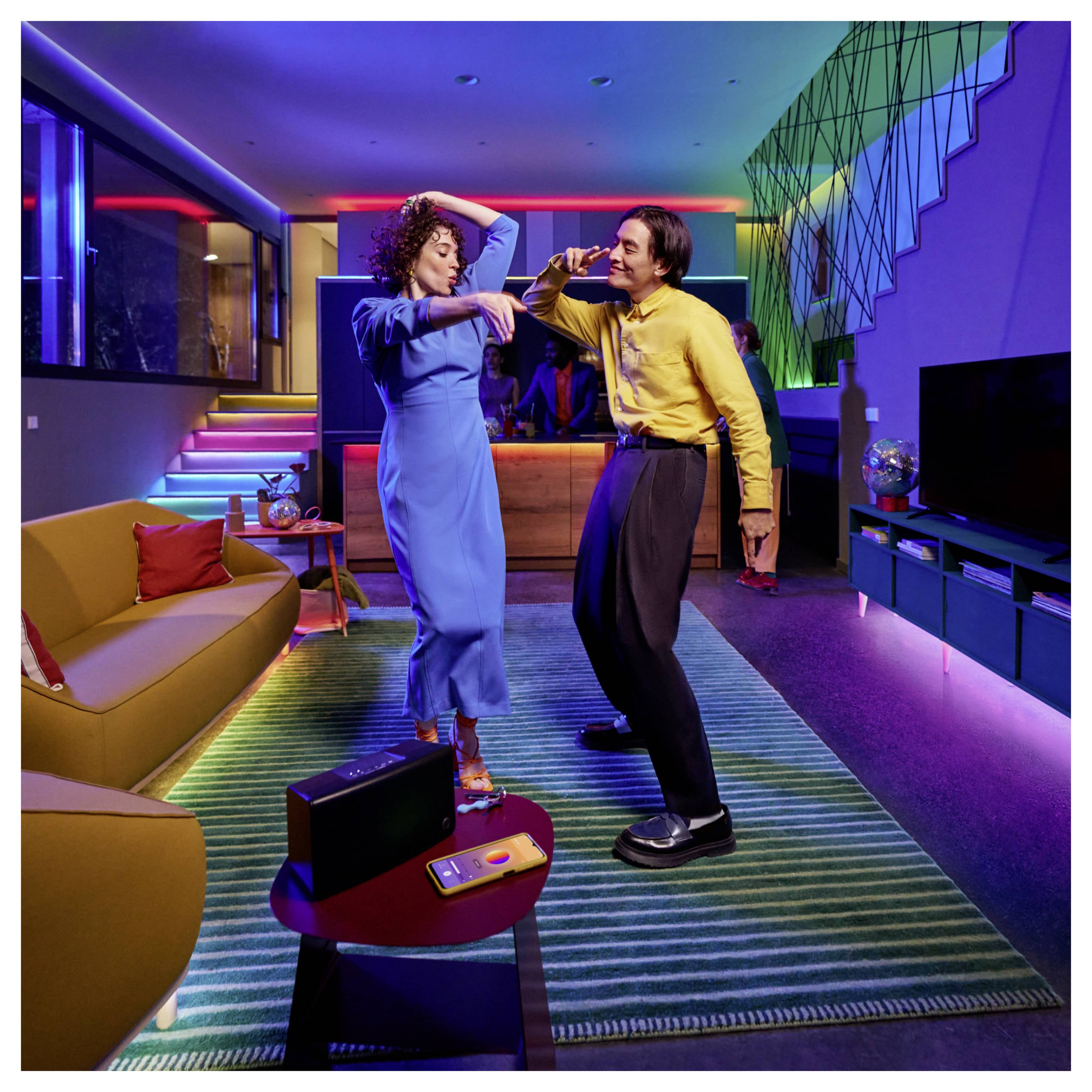 A man and woman dance energetically in a colorful, modern living room with bright, multicolored lighting and stylish decor.