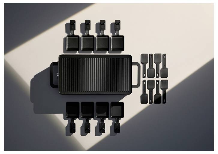 A black raclette grill set arranged with eight small pans, spatulas, and a central rectangular grill. It is lit by sunlight from the left.