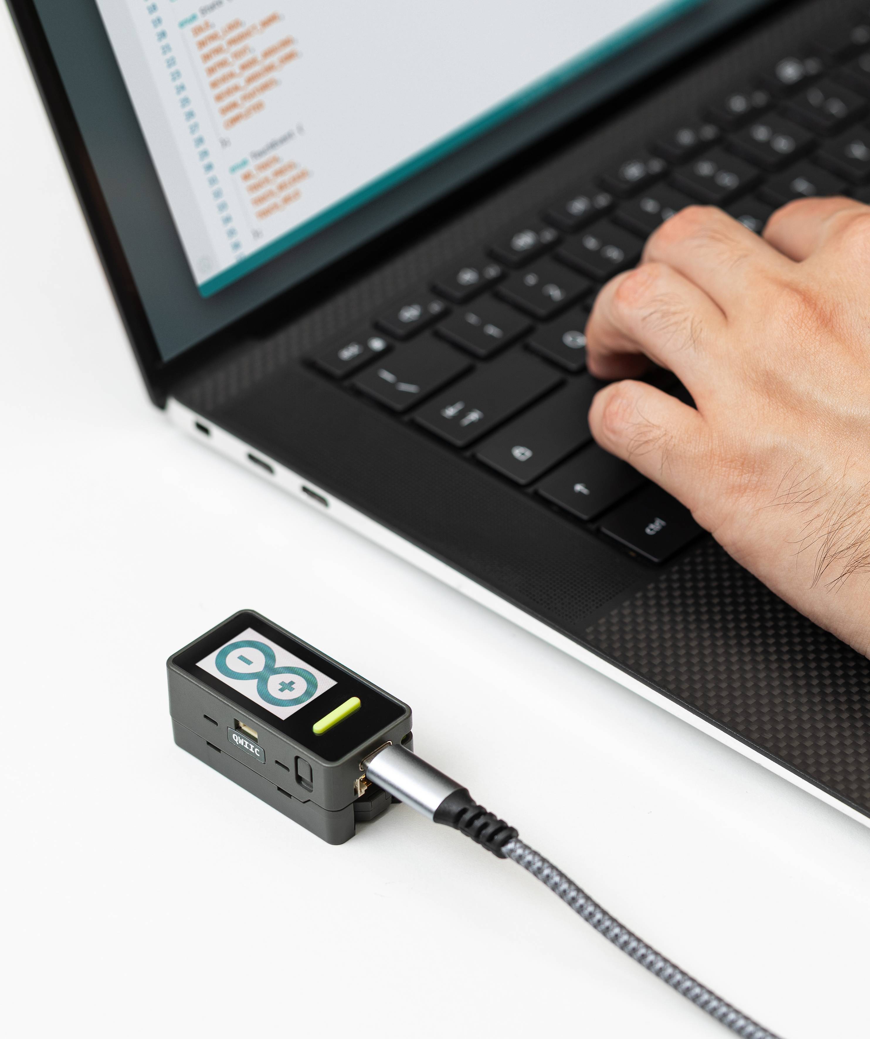 'Person typing on a laptop with coding software on the screen, connected to an Arduino board via a USB cable.'