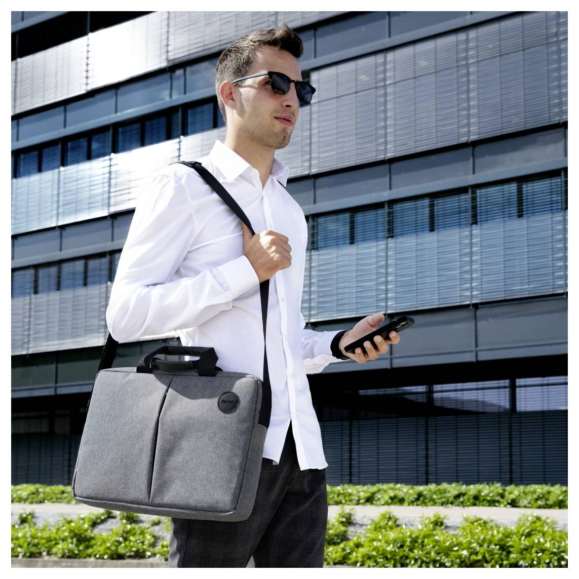 A person in sunglasses and a white shirt walks outside a modern building, holding a gray briefcase and looking at a smartphone.