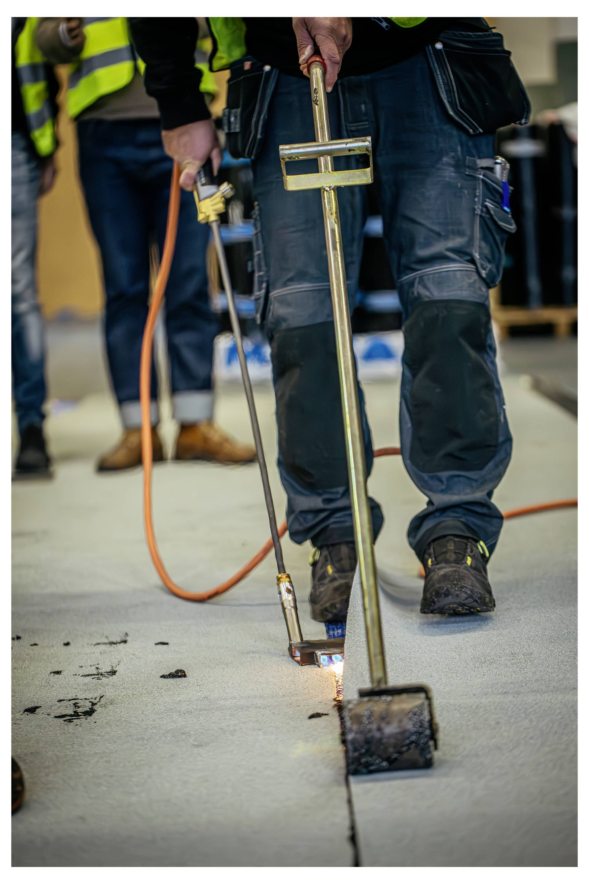 A worker in protective clothing uses a blowtorch to seal a roofing material seam. Safety equipment and other workers are in the background.