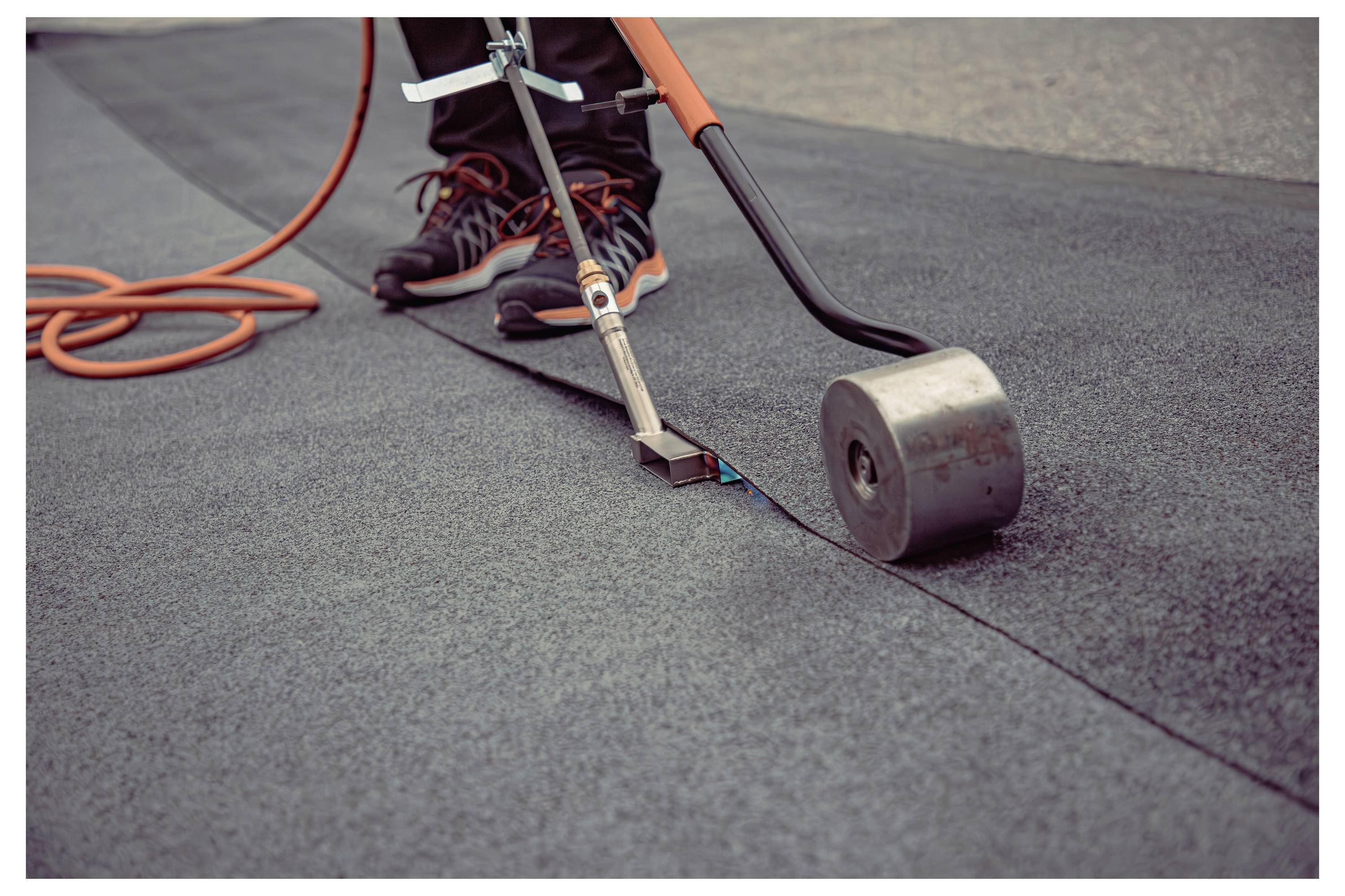 A person using a torch and roller to seal roofing material on a flat surface. The image shows an orange hose and detailed footwork.