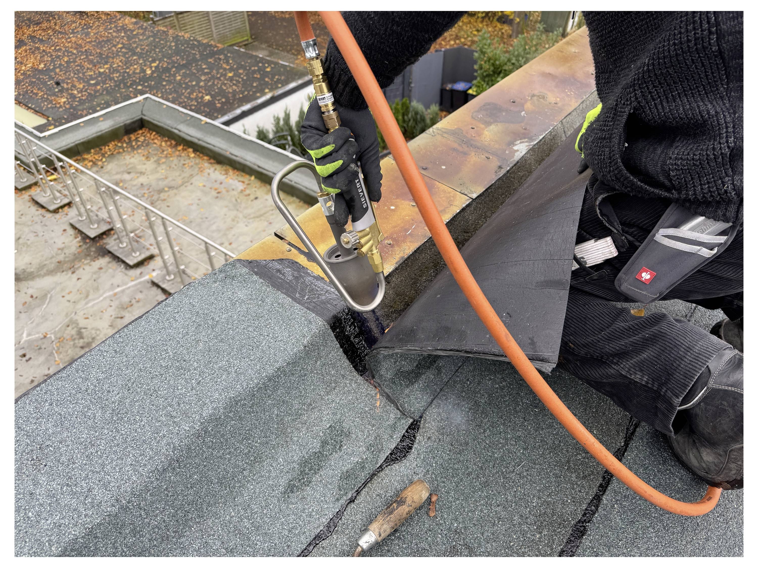 A person is kneeling on a rooftop, using a gas torch to seal roofing material. Safety equipment is visible, and autumn leaves can be seen.