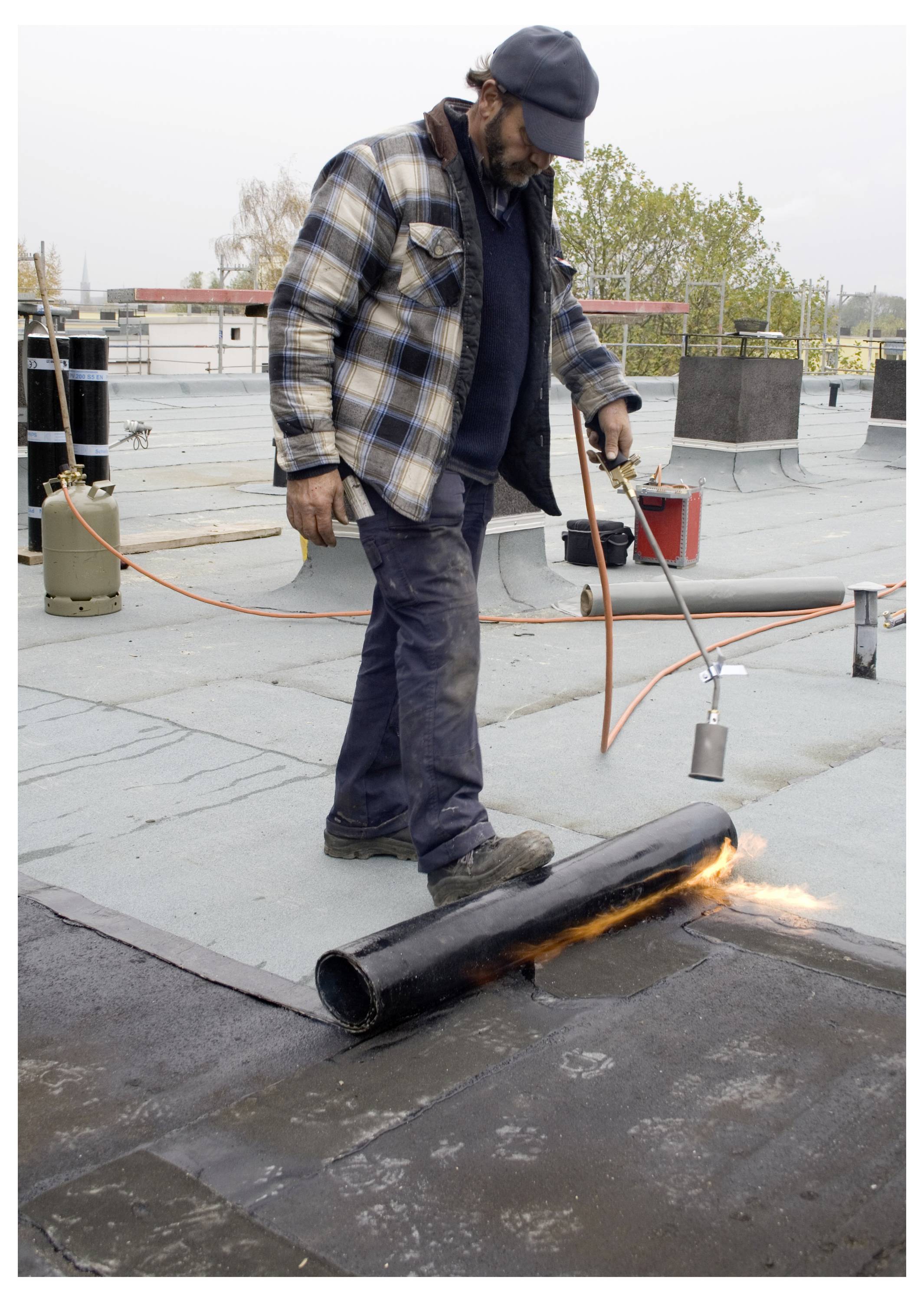 A worker wearing a plaid jacket and cap uses a torch to melt and apply roofing material on a flat roof. Equipment is scattered around.