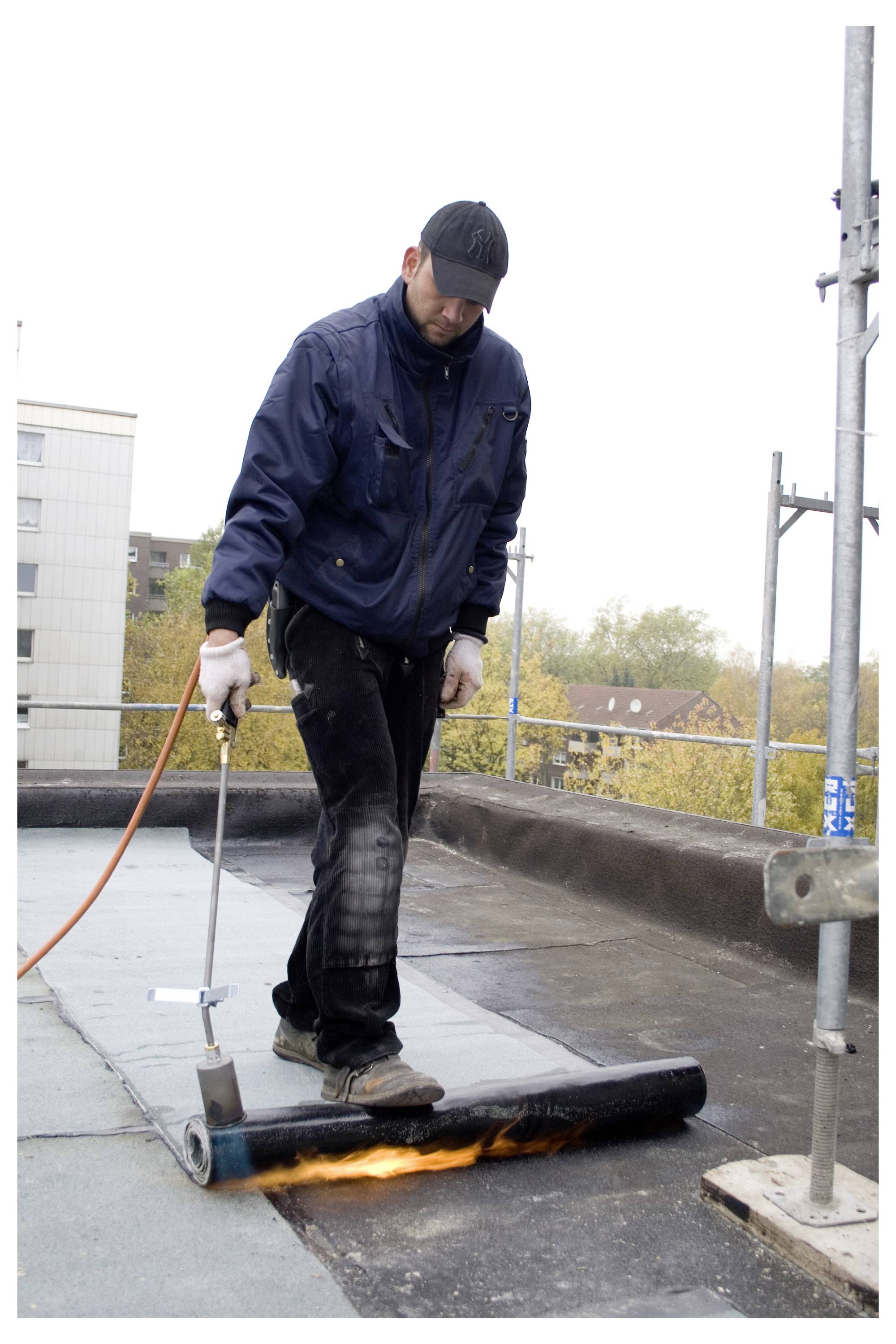 A worker wearing protective clothing applies a torch to a roofing material on a flat rooftop, sealing the surface with heat.