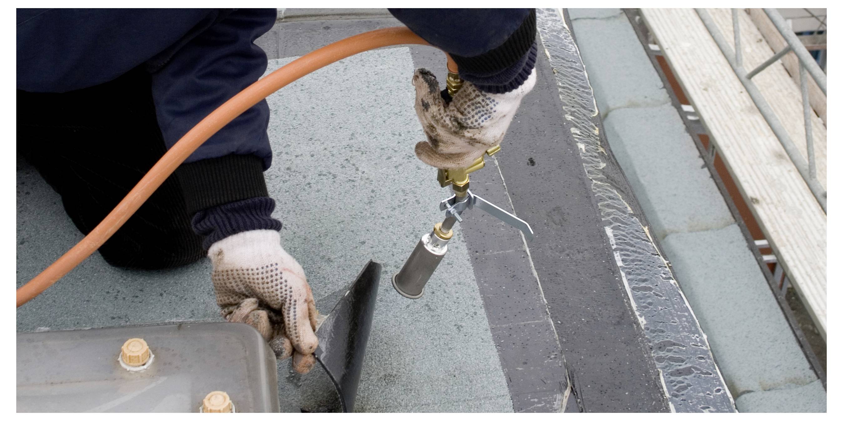 A worker wearing gloves uses a hot air gun to seal roofing material on a flat roof, with an orange hose connected to the tool.